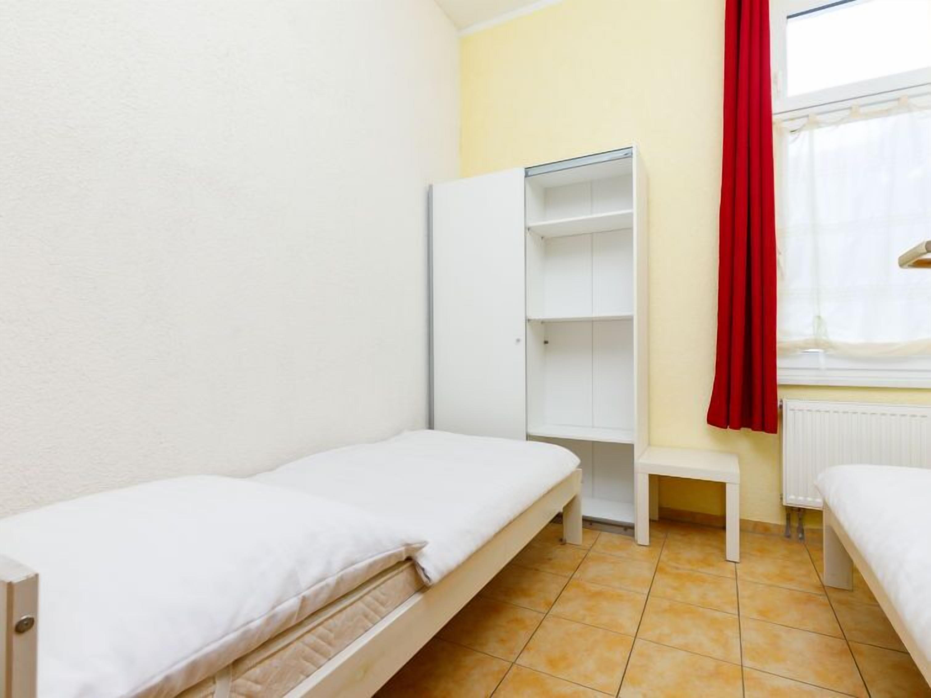 Un pequeño dormitorio individual con dos camas, un armario blanco, una mesita y una ventana con cortina roja.