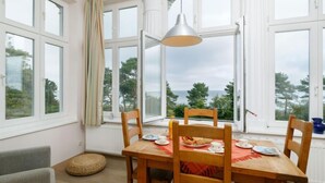 A dining area with a wooden table, four chairs, and a hanging pendant light. Large windows with white frames offer views of trees and the sky. - App.6 Zinnowitzer Hof by Interhome (Zinnowitz)