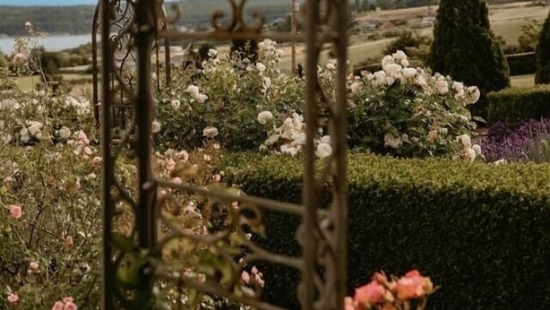 Una reja de hierro forjado da acceso a un jardín con diversas flores y vistas al paisaje.