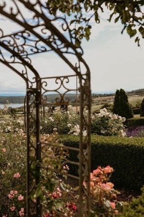 A wrought iron gate leading to a garden with various flowers and a view of the landscape.