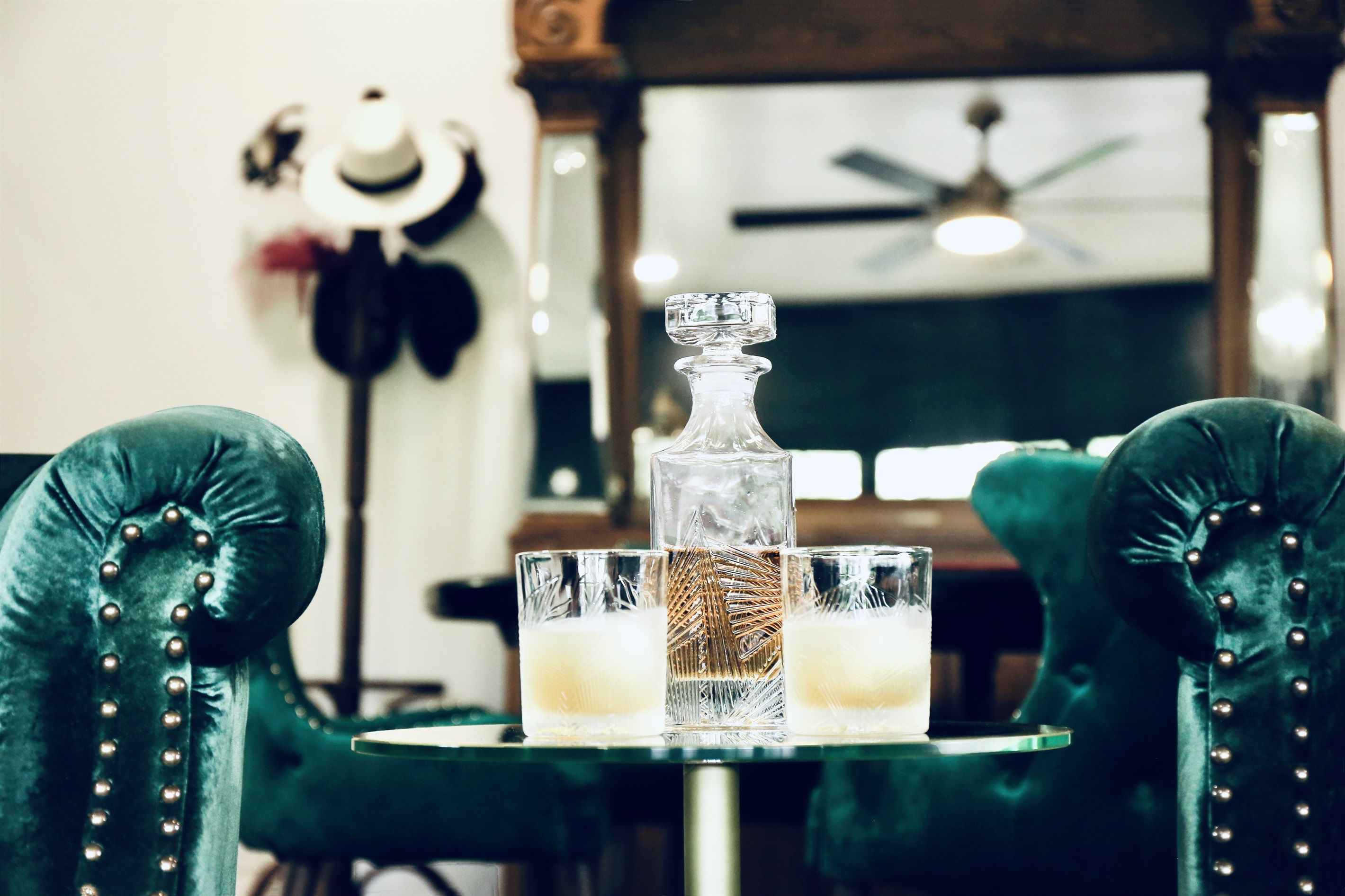 A table with a decanter and two glasses of a light-colored beverage, set between two upholstered chairs with button details.