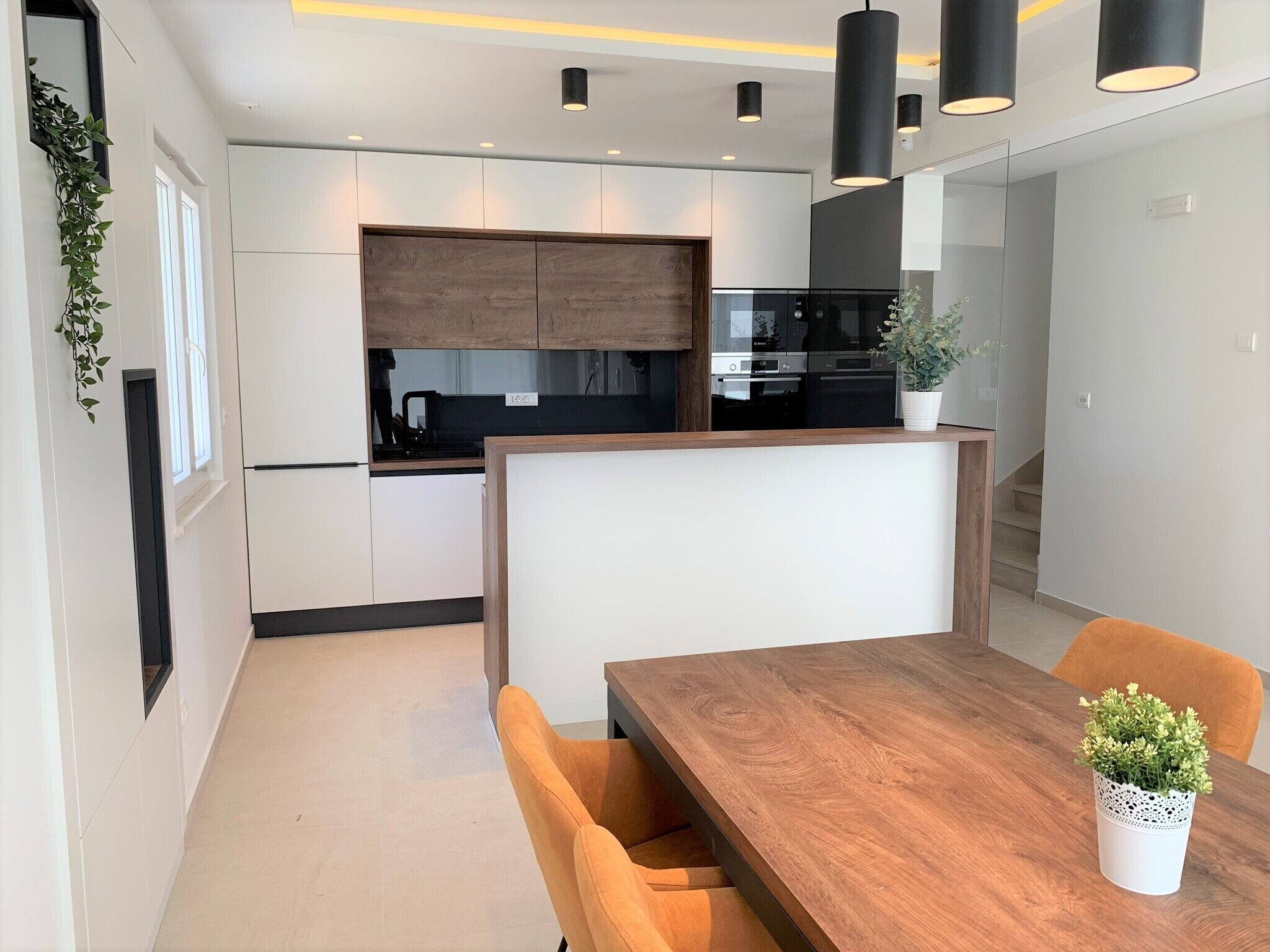 A modern kitchen with white cabinetry, a wooden countertop, and black appliances. There is a dining area with a wooden table and orange chairs.