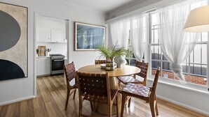 Een eetgedeelte met een ronde houten tafel, zes geweven stoelen, een vaas met groene planten en een groot raam met dunne gordijnen.