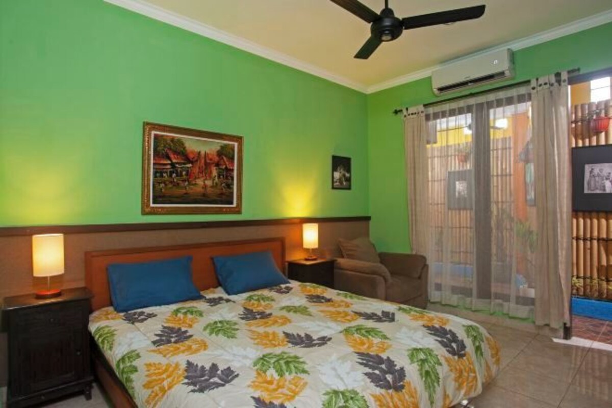 A hotel room with a bed, two bedside lamps, a ceiling fan, a painting on the wall, and a window with curtains.