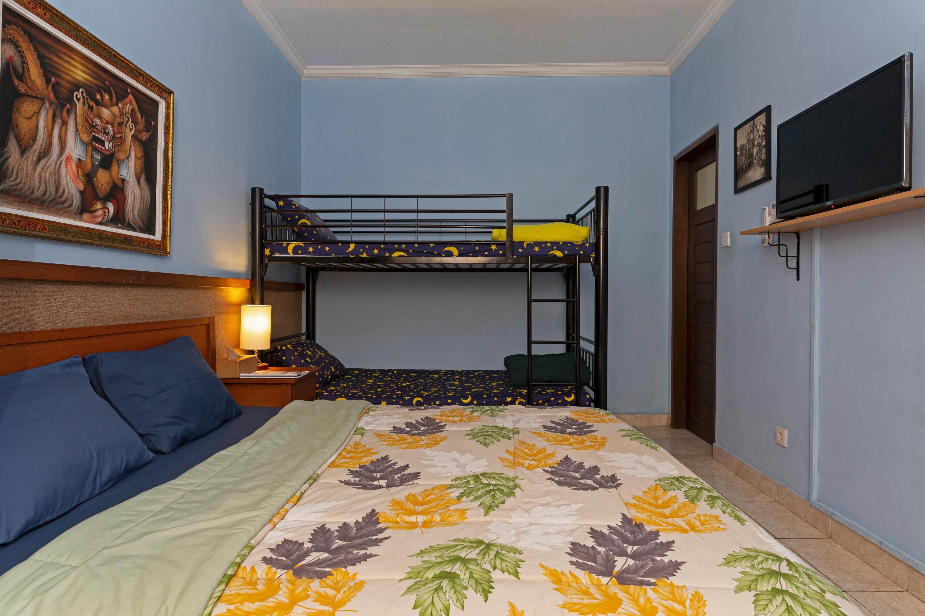Una habitación con una litera, una cama con colcha estampada con motivos de hojas, una mesita de noche con lámpara, un cuadro enmarcado en la pared y un televisor en un estante.