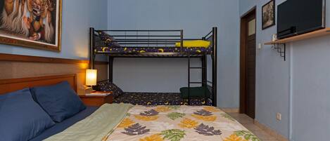 Una habitación con una litera, una cama con colcha estampada con motivos de hojas, una mesita de noche con lámpara, un cuadro enmarcado en la pared y un televisor en un estante.