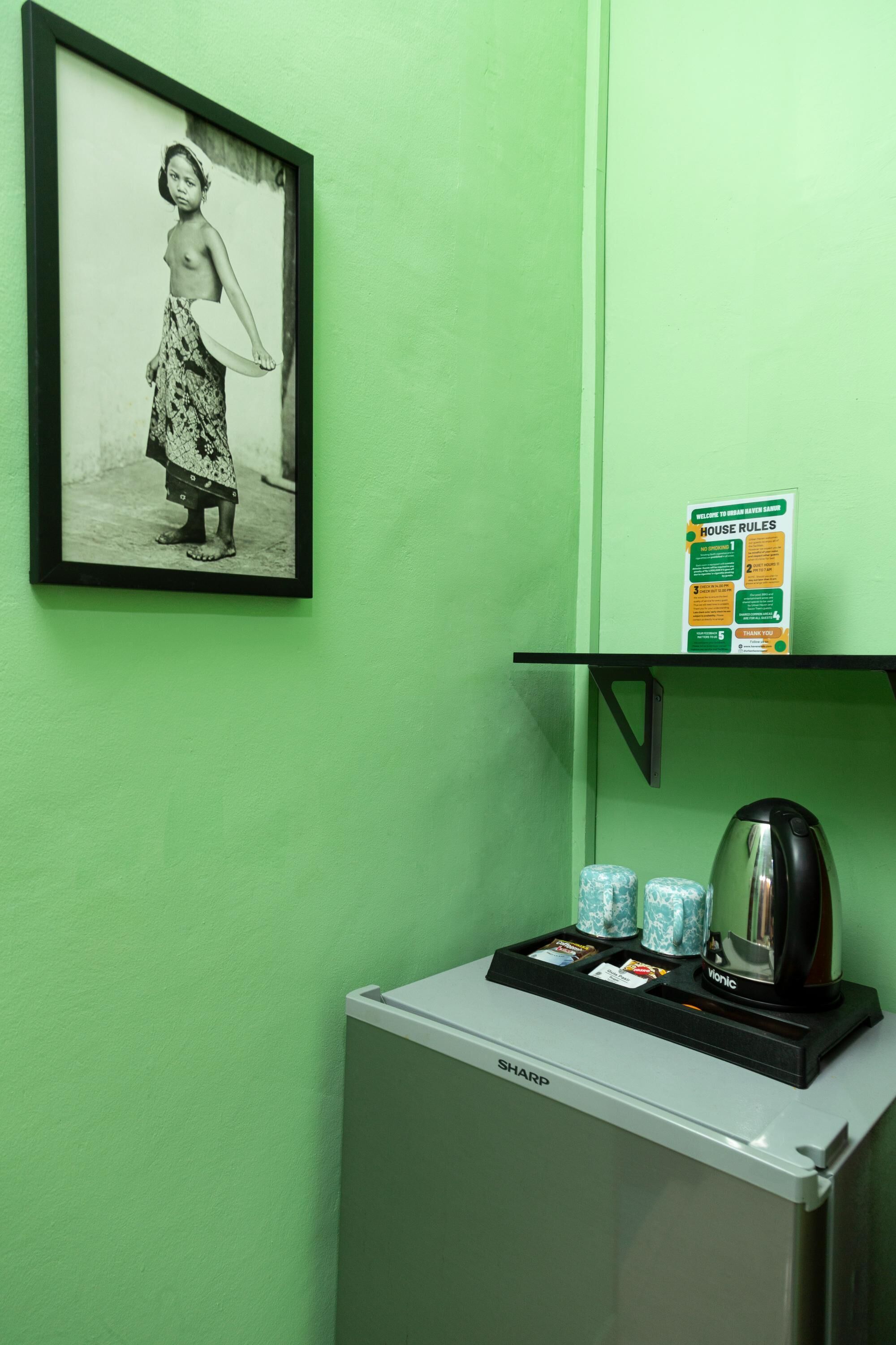 A room with a green wall, a framed black and white photograph, a coffee machine, and a small shelf with a house rules sign.