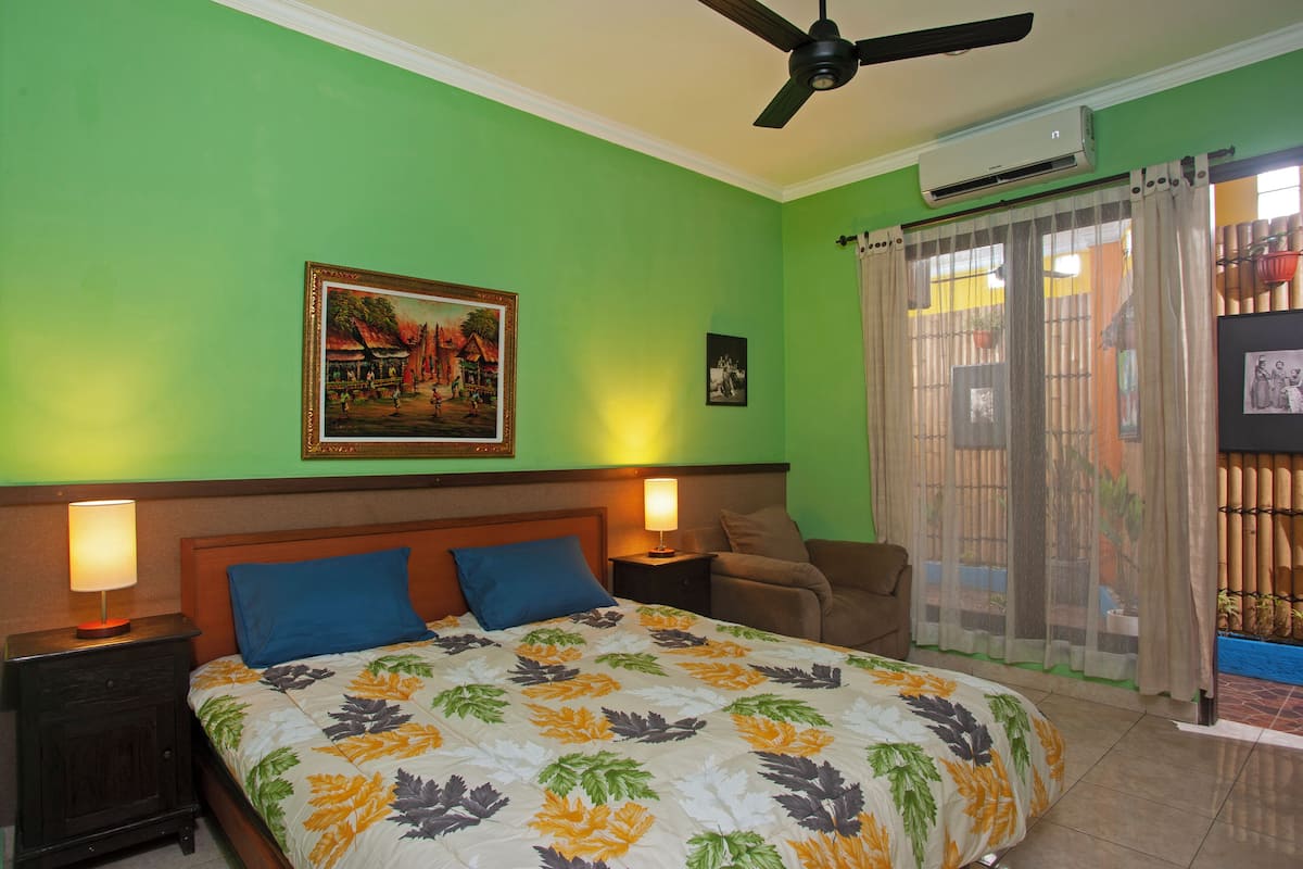 A bedroom with a bed, two nightstands, a ceiling fan, an air conditioning unit, a window with curtains, and a wall-mounted picture.
