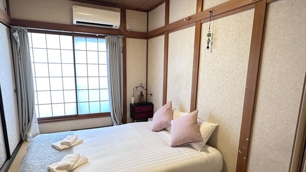 A hotel room with a bed, pillows, a nightstand, a window with curtains, and an air conditioning unit. - Tabi Time Oimazatominami (Osaka)