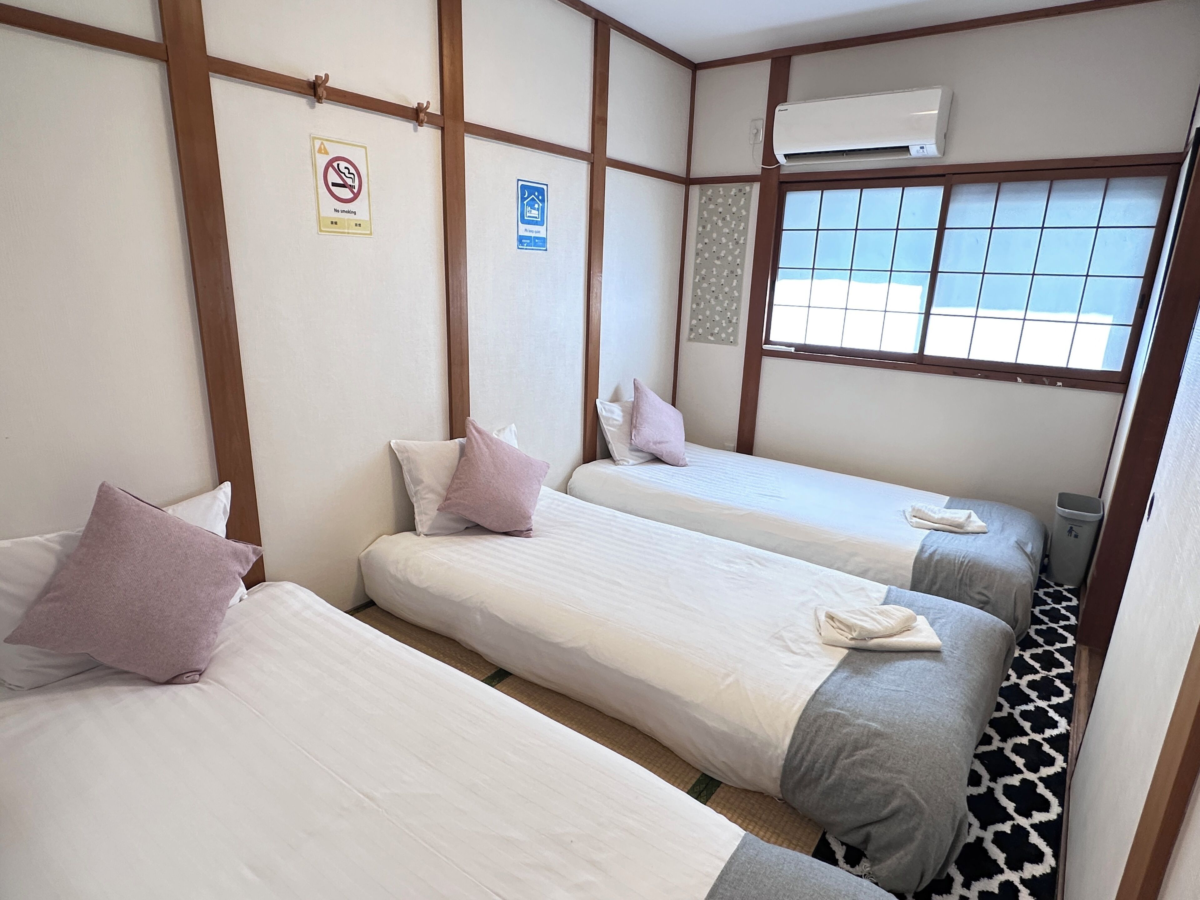 Une chambre d’hôtel avec trois lits, un panneau indiquant qu’il est interdit de fumer, un panneau bleu signalant qu’il s’agit d’une chambre privée, et un appareil de climatisation.