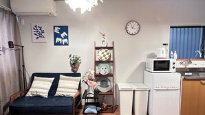 A compact living space with a sofa, a small table with a chair, a decorative shelf, a clock, and a kitchenette area. - Tabi Time Oimazatominami (Osaka)