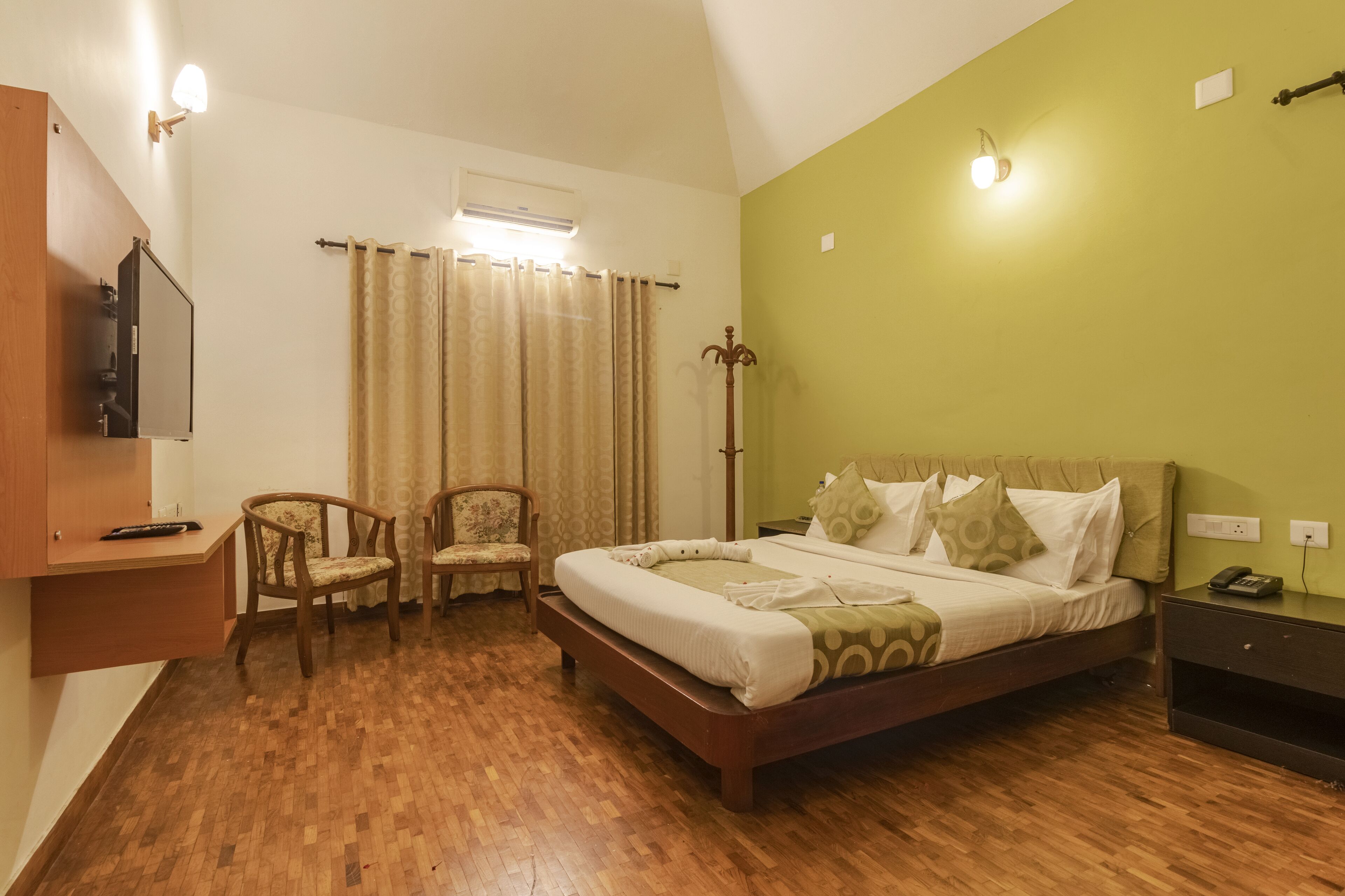 A bedroom with a wooden floor, a bed with patterned bedding, a chair, a bedside table with a phone, and a wall-mounted light fixture.