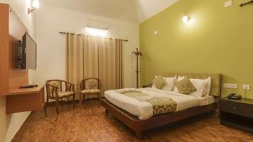 A bedroom with a wooden floor, a bed with patterned bedding, a chair, a bedside table with a phone, and a wall-mounted light fixture.