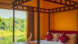 Uma cama com dossel adornada com enfeite de cisne, uma poltrona florida e vista para um lago através de amplas janelas.