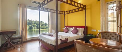A four-poster bed with a wooden frame, a wooden table, and a chair in a room with yellow walls and a large window overlooking a lake.