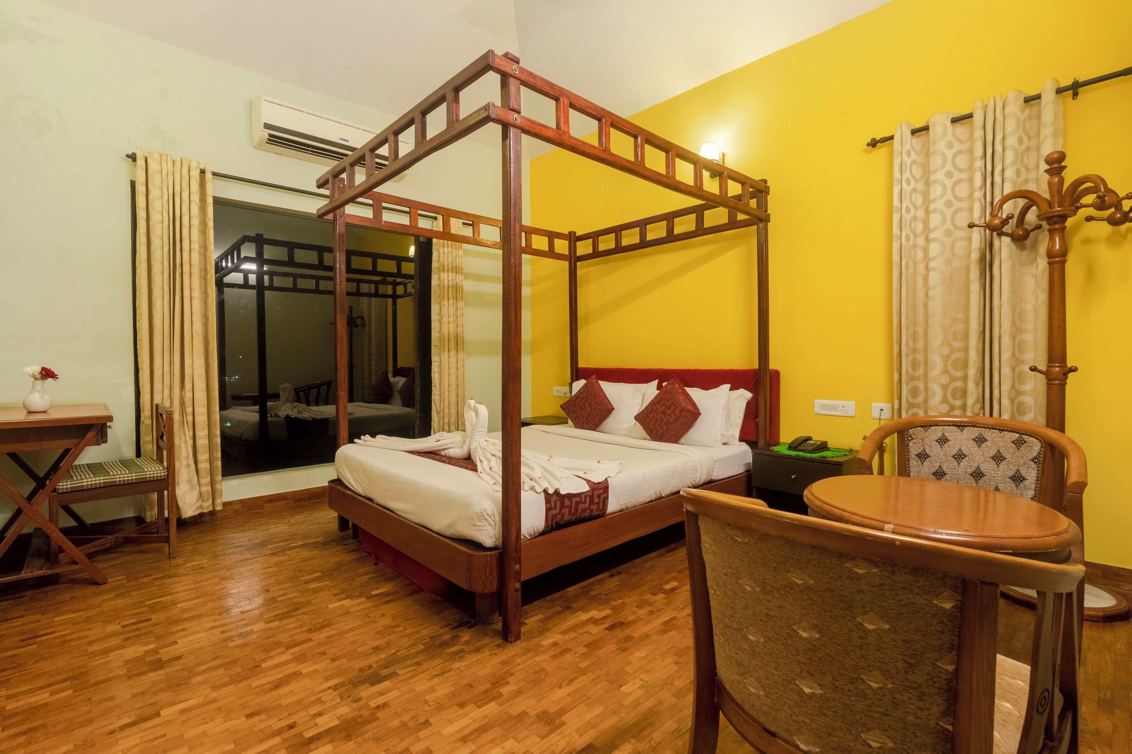 A four-poster bed with white linens, a wooden floor, a dining table, and a chair.