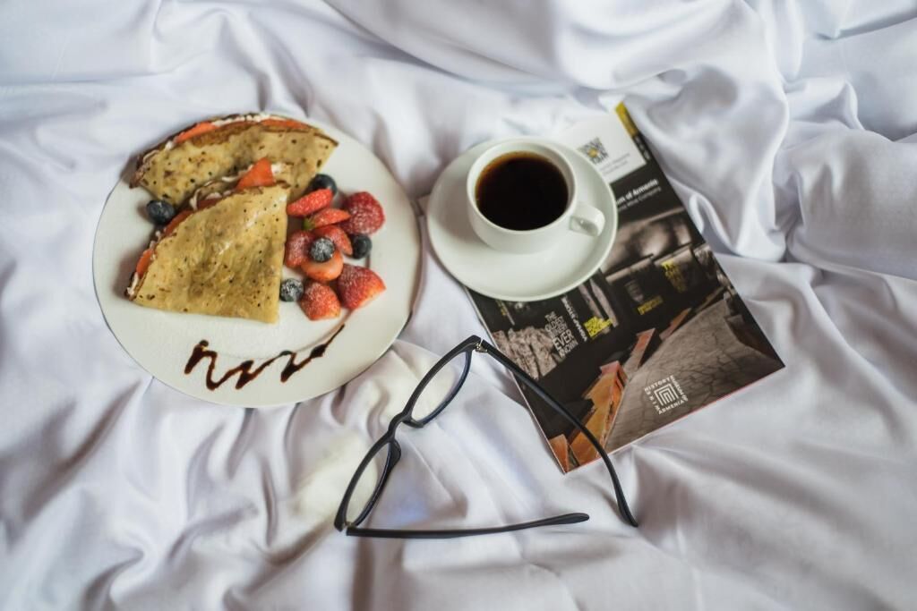 Un plato con crepas, fresas y arándanos, una taza de café y una revista sobre un edredón blanco.