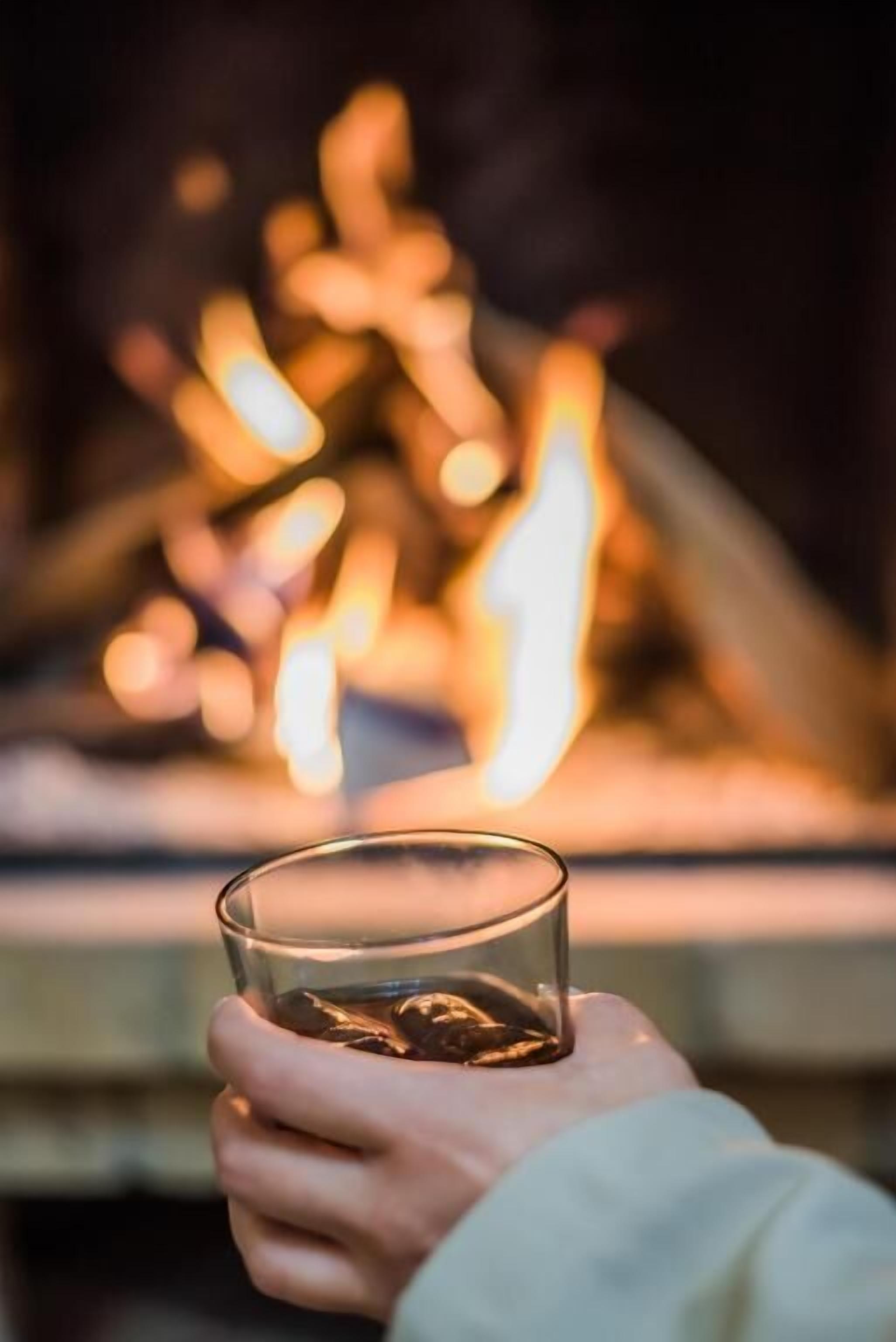 Een hand houdt een glas met vloeistof vast, voor een onscherpe open haard.
