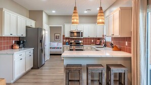A modern kitchen with white cabinets, a central island with stools, a refrigerator, a stove, and hanging pendant lights. - Hacienda San Miguel | Resort Style Pool W/ Sunken Outdoor Kitchen (Indio)