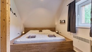 Una camera da letto con soffitto spiovente, un letto con testiera in legno, un comodino con una lampada, una finestra con tende e una mensola a parete con una bicicletta e un quadro.