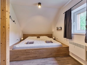 A bedroom with a sloped ceiling, a bed with a wooden headboard, a bedside table with a lamp, a window with curtains, and a wall-mounted shelf with a bicycle and picture. - Vacation home Muhu by Interhome (Smržovka)