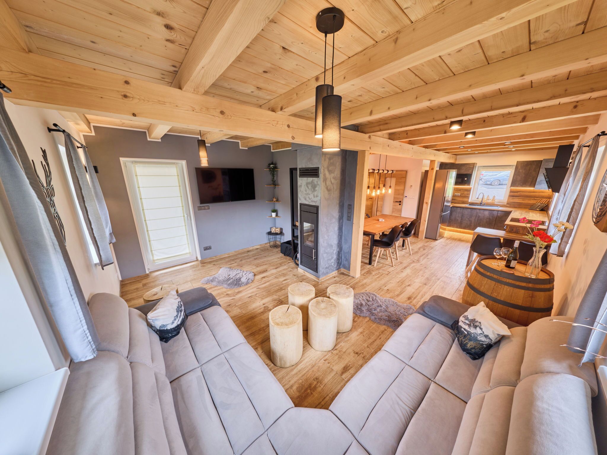 Un interior moderno con techo de madera, un área de descanso con sofá y reposapiés, una zona de comedor con mesa y sillas, y una cocina visible al fondo.