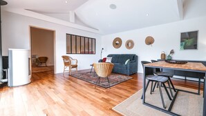 Ein modernes Wohnzimmer mit Holzboden, einem grauen Sofa, Korbstühlen, einem Esstisch mit schwarzen Stühlen und einem weißen Kühlschrank.