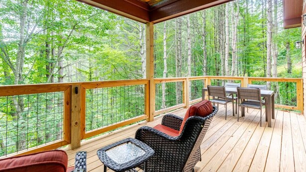 A covered deck with wicker furniture and a table, overlooking a forested area. - Spring in Mtns Poplar Treehouse Near Asheville - Pet Friendly (Marshall)