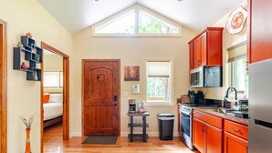 A compact kitchen with wooden cabinets, a microwave, and a stove. There is a window with blinds, a door, and a small table with a coffee maker. - Spring in Mtns Poplar Treehouse Near Asheville - Pet Friendly (Marshall)
