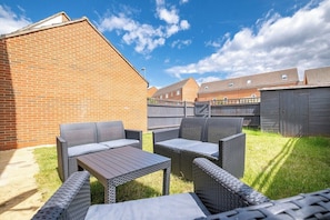 Ein Garten mit Backsteinmauer, Holzzaun und einer Terrasse mit Korbmöbeln und einem Tisch.