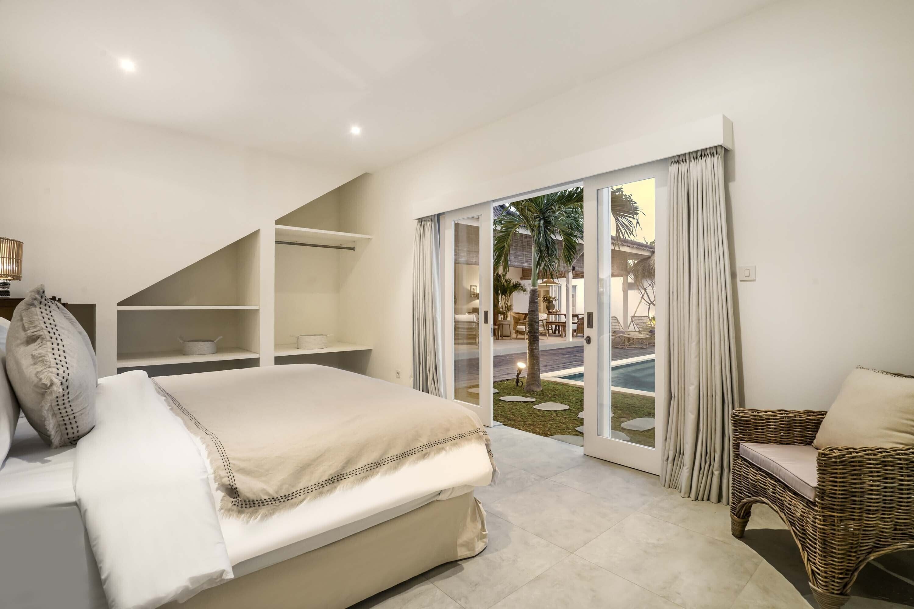 A bedroom with a large bed, a wicker chair, and a view of a pool through glass doors.