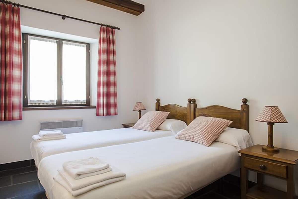 Habitación de hotel con dos camas, una ventana con cortinas, una mesita de noche con lámpara y un radiador.
