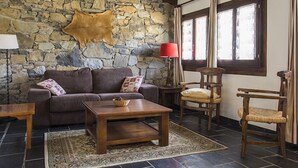 A cozy living room with a stone wall, a leather hide, a brown sofa, a wooden coffee table, a red floor lamp, and a window with curtains. - Apartamentos Linsoles (Benasque)