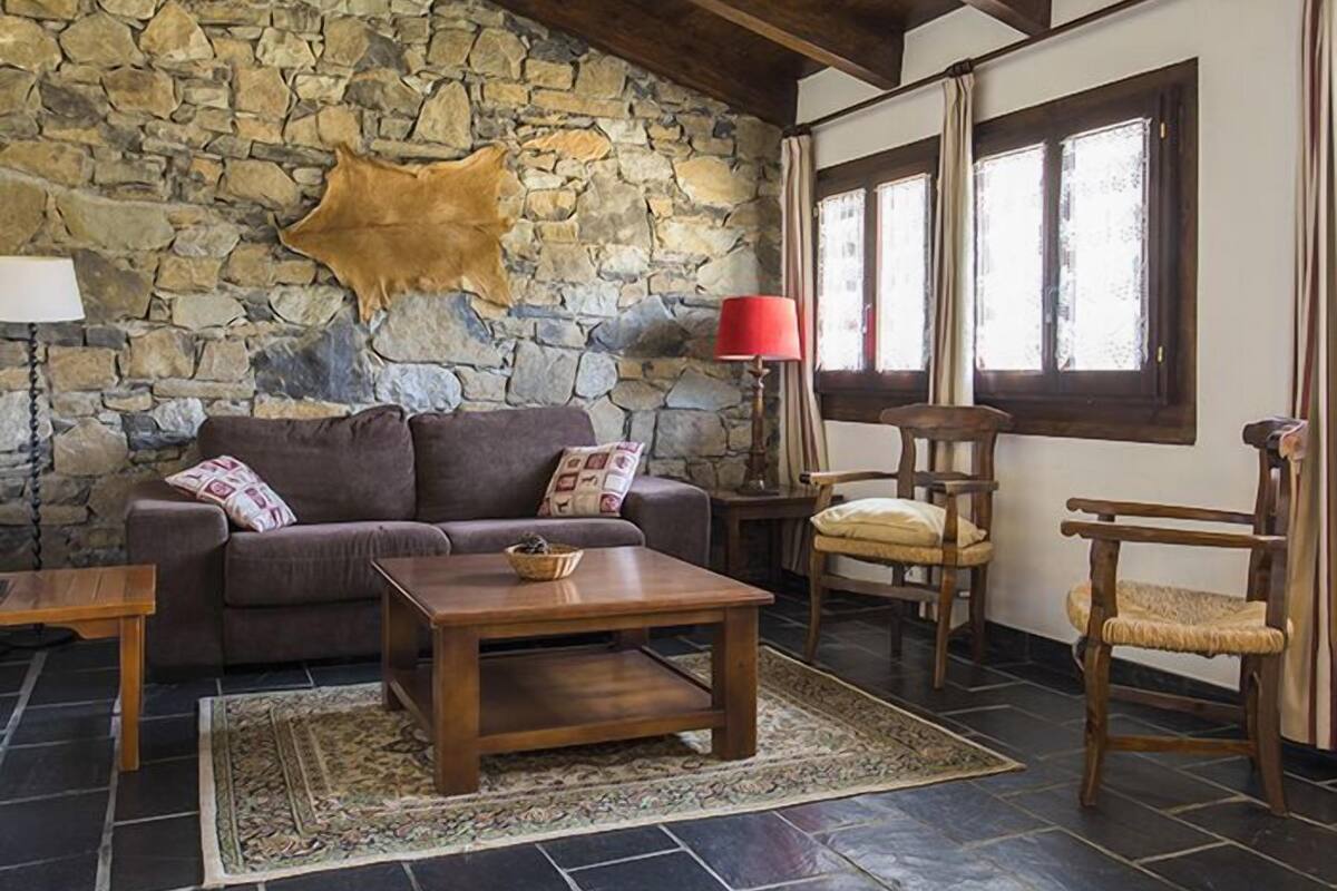 Una sala de estar acogedora con pared de piedra, un cuero, un sofá marrón, una mesa de centro de madera, una lámpara de pie roja y una ventana con cortinas.