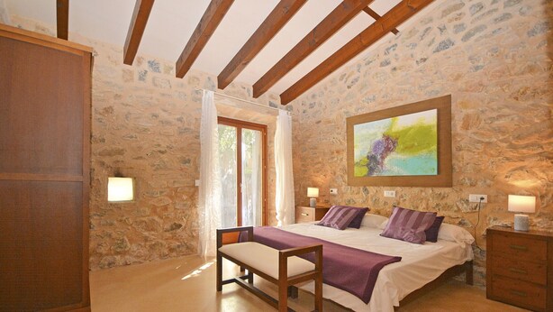 A bedroom with a stone wall, wooden beams, a bed with a purple blanket, a bench, and a painting. - Es Romagueral - Beautiful house with forest, pool, and air conditioning in Mallorca (Arta)