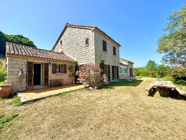 Une maison en pierre avec un toit de tuiles, un banc en bois et une table de pique-nique dans une cour verdoyante.