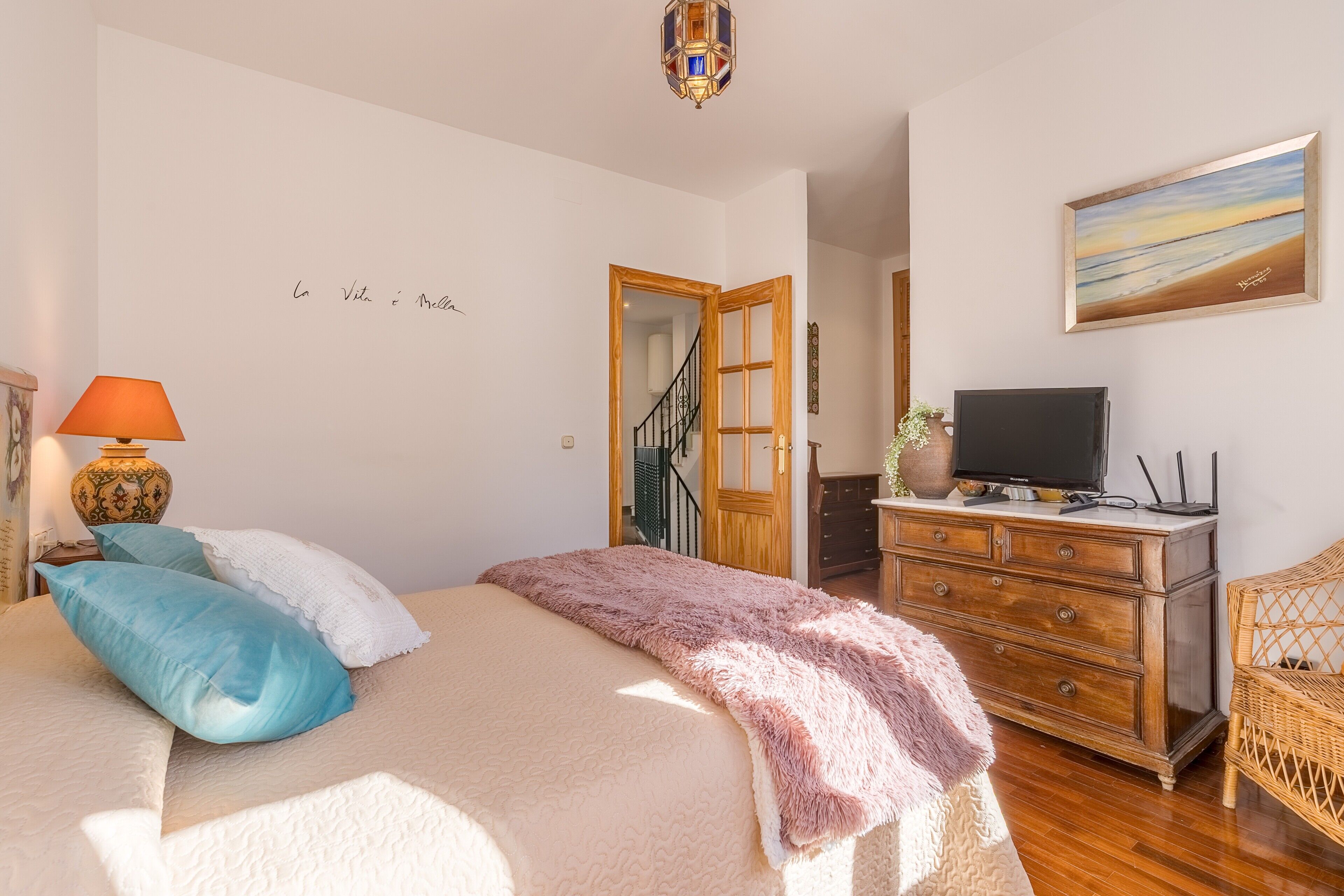 A bedroom with a bed, a television on a dresser, a painting on the wall, and a staircase leading to another room.