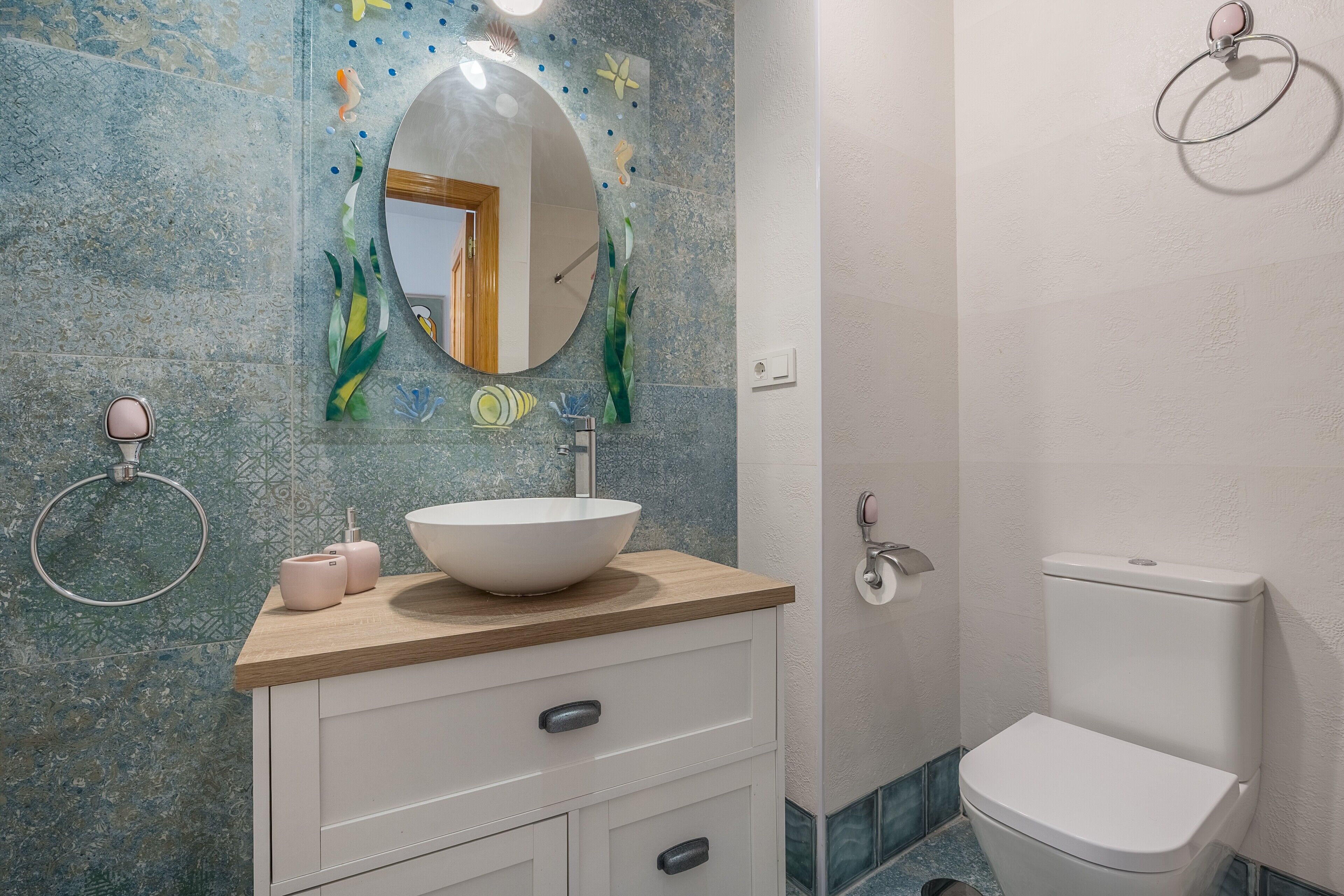 Une salle de bain avec un miroir rond, un lavabo blanc, des toilettes et des carreaux décoratifs sur les murs.