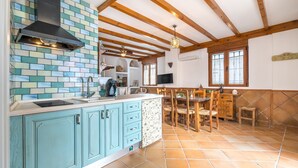 A kitchen with blue cabinets, a black range hood, a tiled backsplash, wooden beams, a dining area with a table and chairs, and a TV mounted on the wall. - Vacation Home "El Valle Verde" with Mountain View, Private Terrace & Wi-Fi (Lecrin)