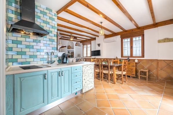 A kitchen with blue cabinets, a black range hood, a tiled backsplash, wooden beams, a dining area with a table and chairs, and a TV mounted on the wall. - Vacation Home "El Valle Verde" with Mountain View, Private Terrace & Wi-Fi (Lecrin)