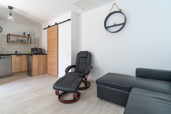 Un salon moderne comprenant un fauteuil inclinable, un canapé, un coin cuisine avec un meuble en bois et une étagère fixée au mur.