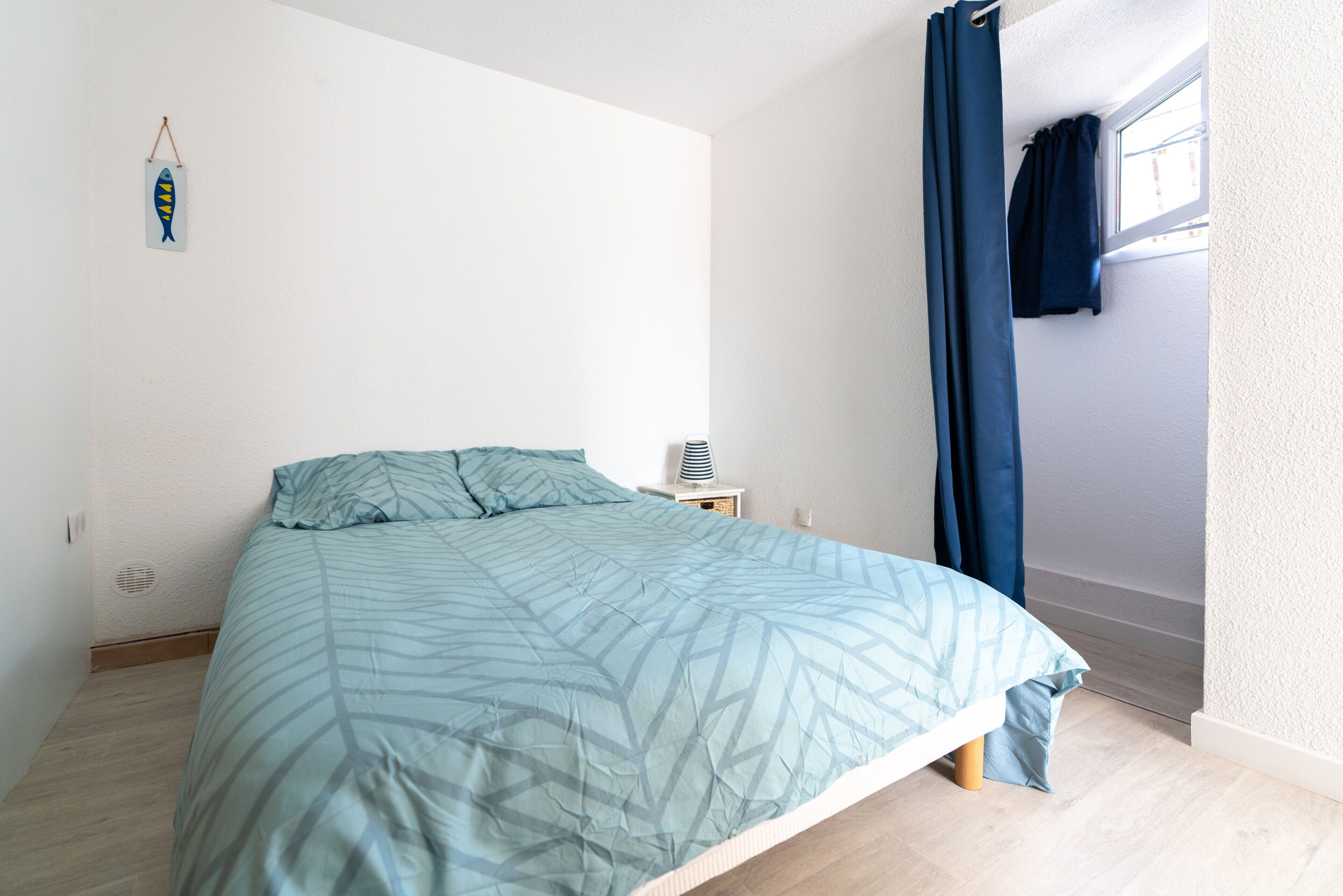 Un dormitorio con cama, mesita de noche, estante en la pared y ventana con cortinas.