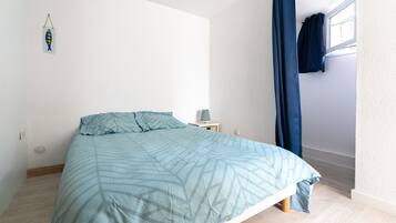 Un dormitorio con cama, mesita de noche, estante en la pared y ventana con cortinas.