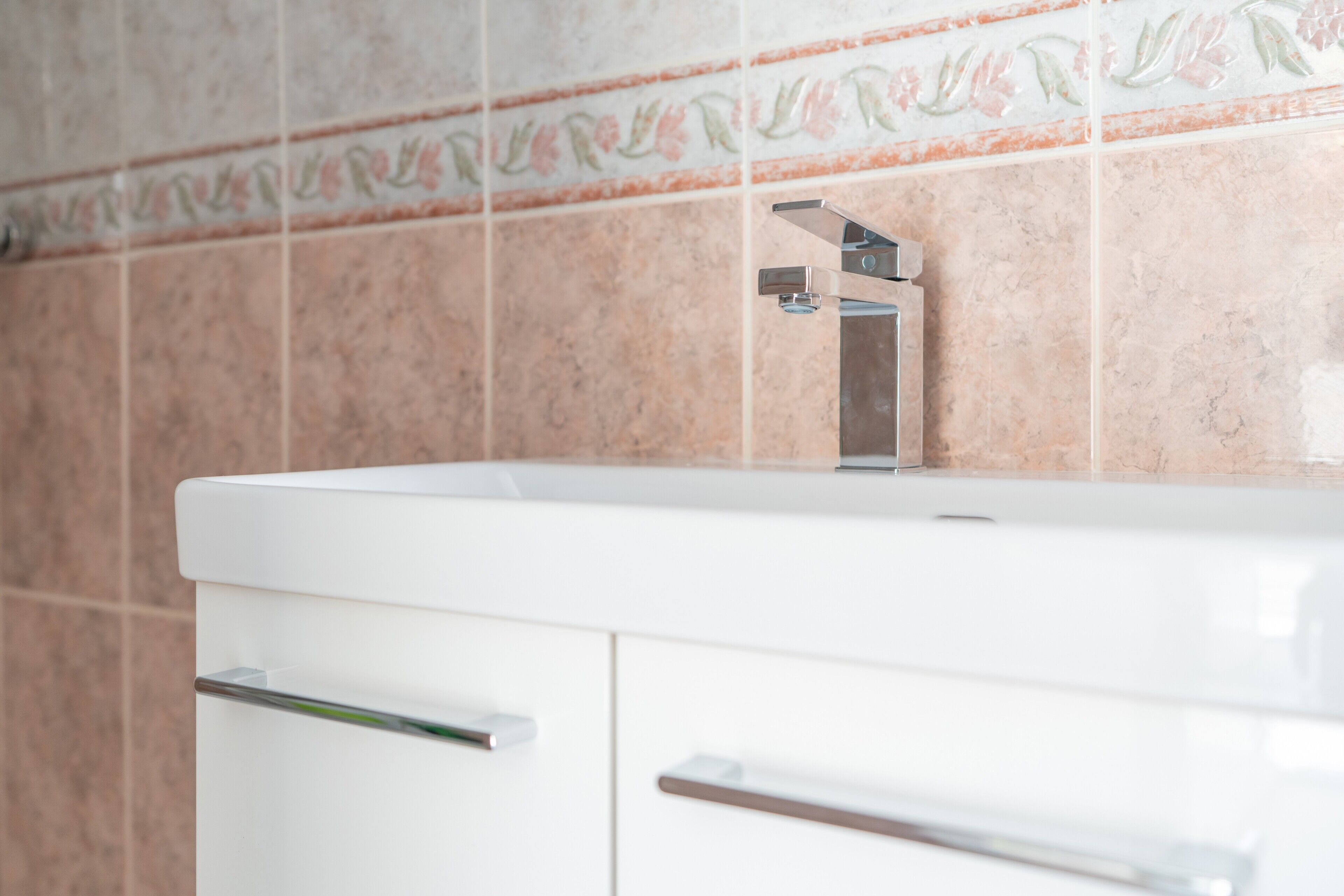 Ein Badezimmer mit einem weißen Waschbecken, einem Chrom-Wasserhahn und einer gefliesten Wand mit einem Blumenmuster.