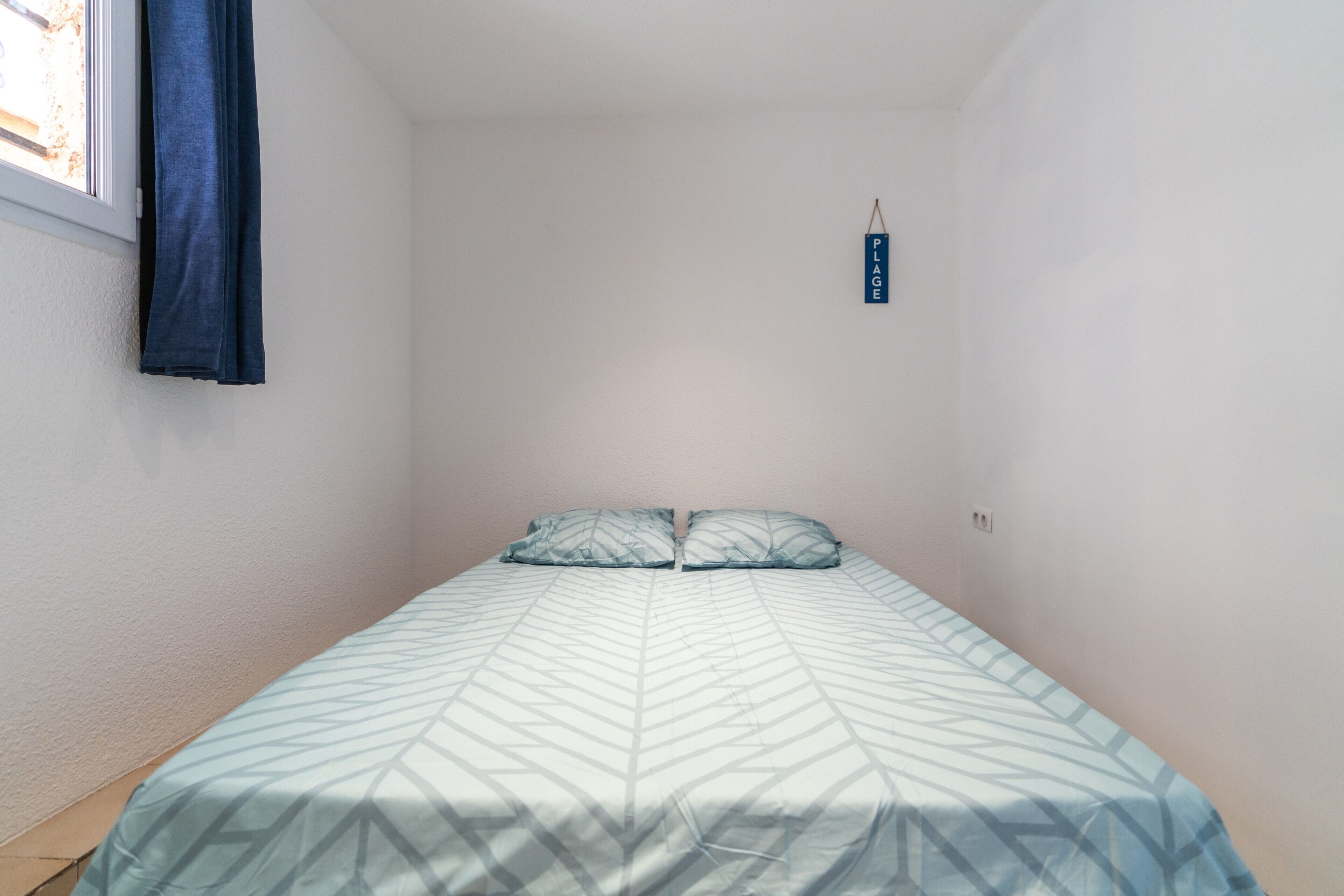 Une petite chambre avec un lit simple, un mur blanc uni, une fenêtre avec un rideau bleu et un petit panneau accroché au mur.