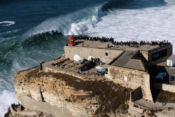 A coastal building with a crowd, a large wave crashing, and a cliff. - Nazaré Sea View Dream Apartment with Ocean View (Nazaré)