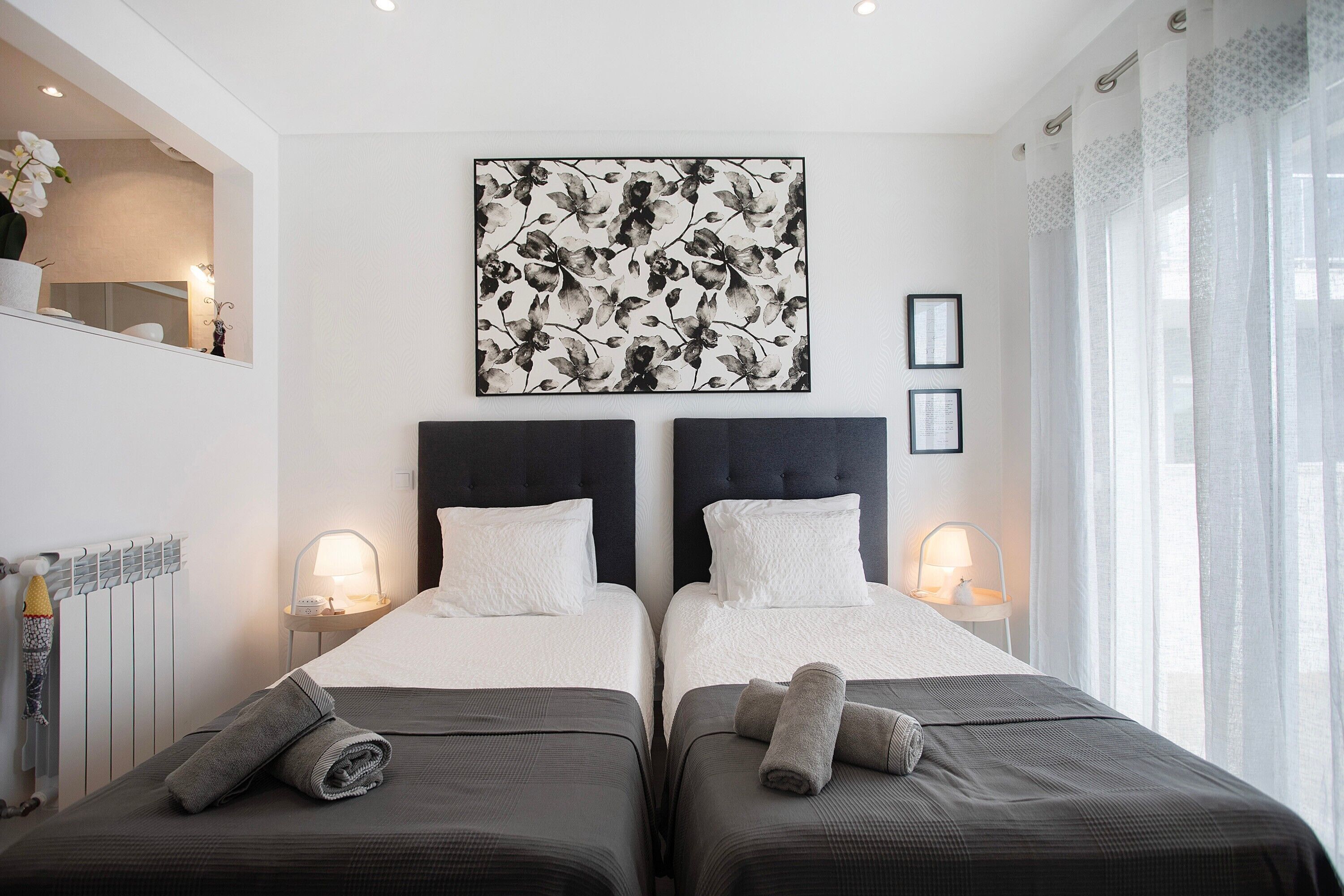 Ein Hotelzimmer mit zwei Betten, einem großen Blumenbild an der Wand, einem Fenster mit transparenten Vorhängen und einem Nachttisch mit Lampe.