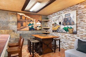 A rustic interior with a wooden table, bar stools, a sofa, and two paintings depicting barrels and wine bottles.