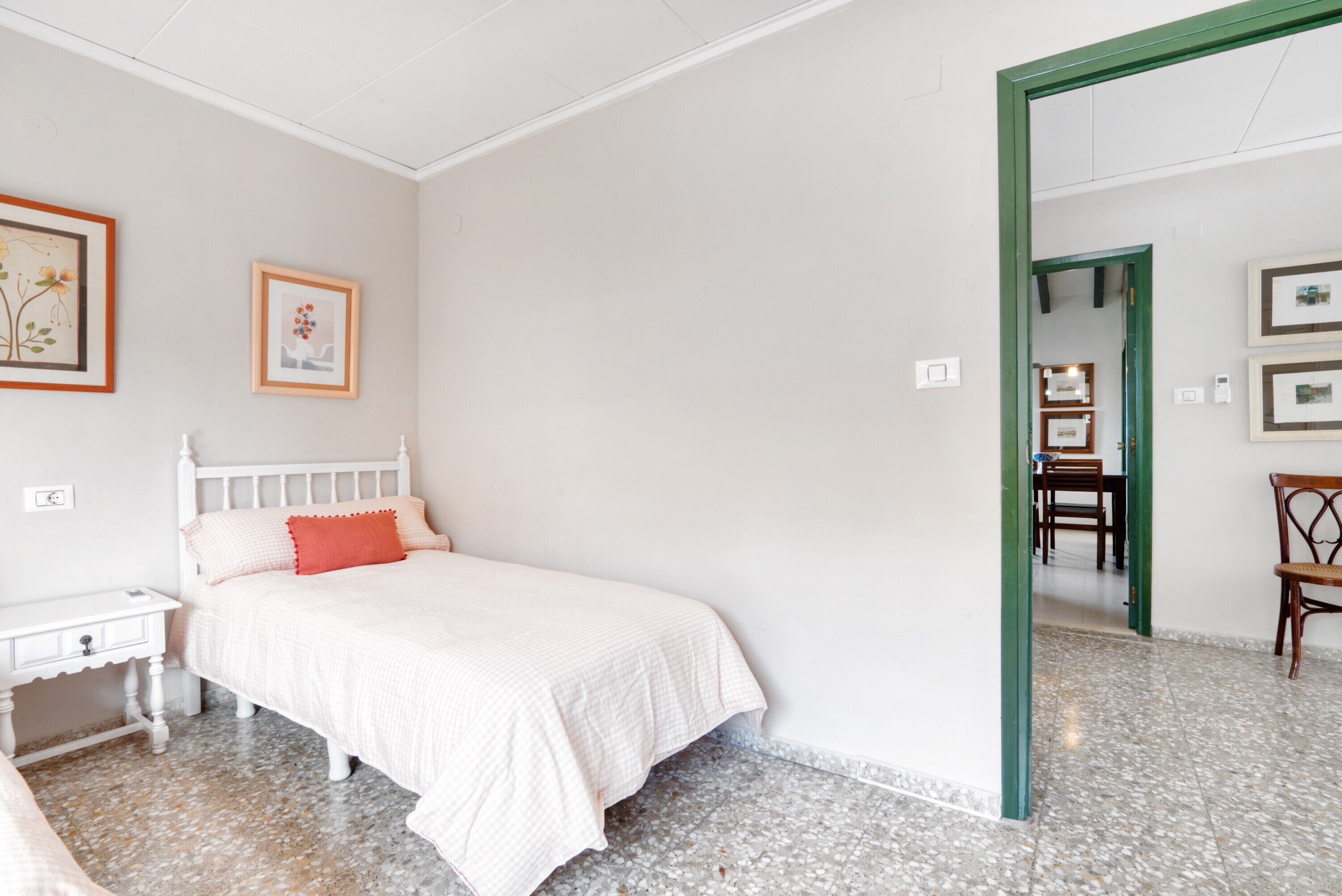 Un dormitorio con cama blanca, una mesita de noche, un cuadro enmarcado en la pared y un vano de puerta verde que da a otra habitación.