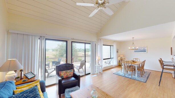 A spacious living room with a large sliding glass door leading to a balcony, a dining area with a chandelier, and a ceiling fan.